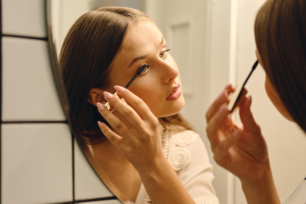 Close up young beautiful woman in white dress standing near mirror and thoughtfully applying mascara in bathroom