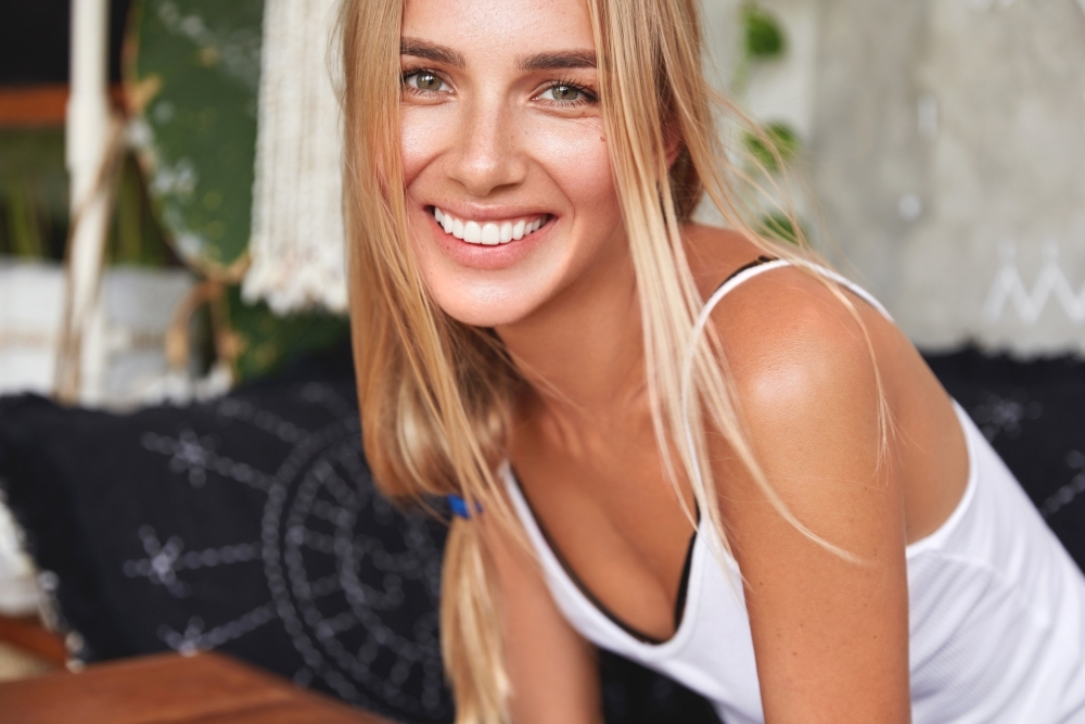 Cute smiling young woman with attractive look has broad smile as laughs joyfully hears positive news from friend have pleasant talk together in cozy domestic atmosphere.