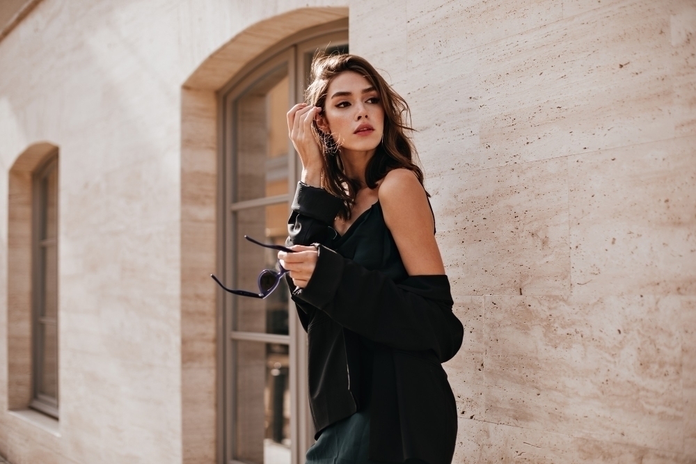 Cute young girl with dark wavy hairstyle and bright makeup silk dress black jacket holding sunglasses in hands and looking away against beige building wall3