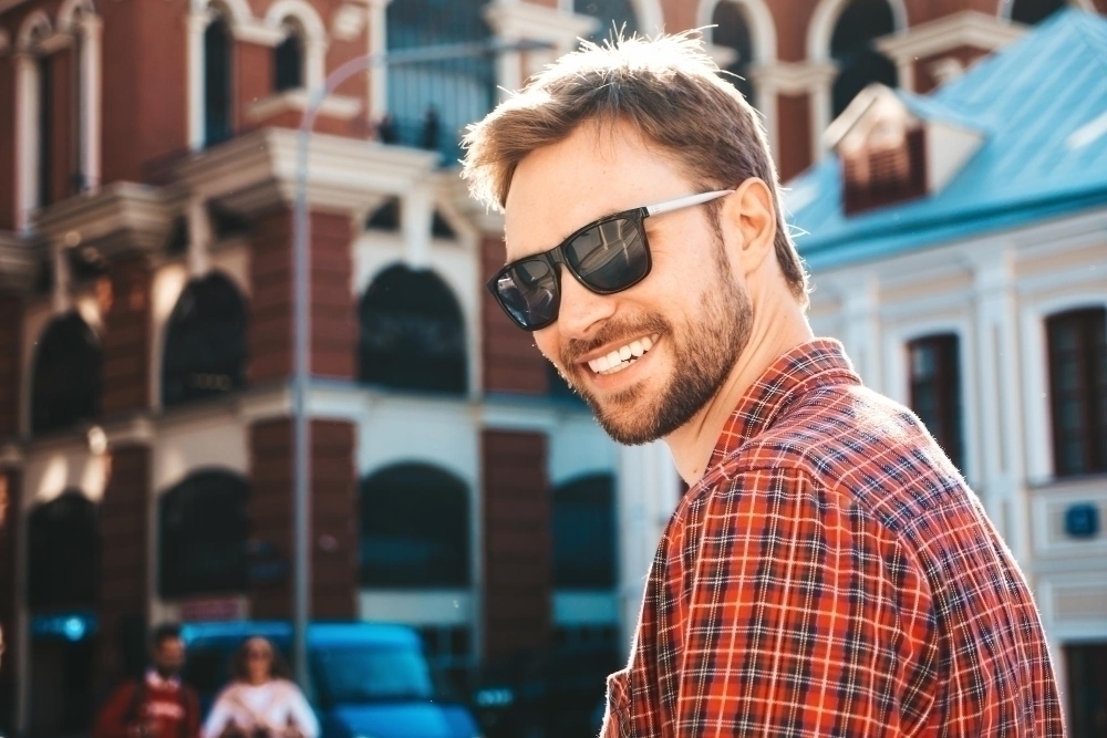 Handsome stylish model.Modern man dressed in red checkered shirt. Fashion male posing near skyscraper on the street background in sunglasses. Outdoors at sunset