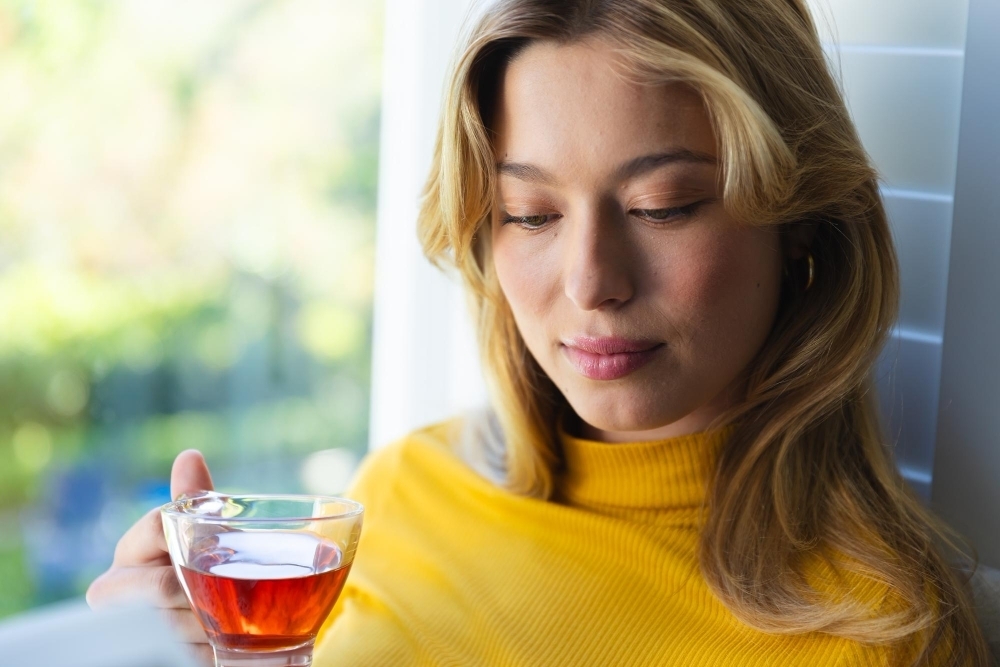 Happy caucasian woman sitting on couch with tea and looking down in sunny room at home