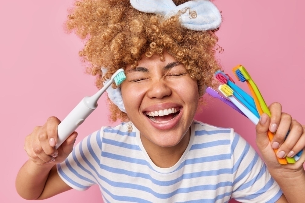 Happy joyful young woman with curly hair holds toothbrushes smiles broadly has white teeth after regular cleaning wears casual striped t shirt keeps eyes closed isolated over pink background