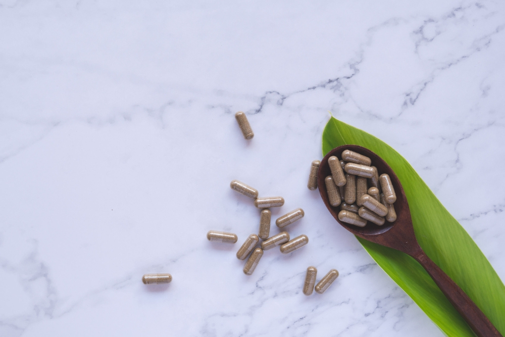 Herbal medicine in capsules on wood spoon with natural green leaf on white marble