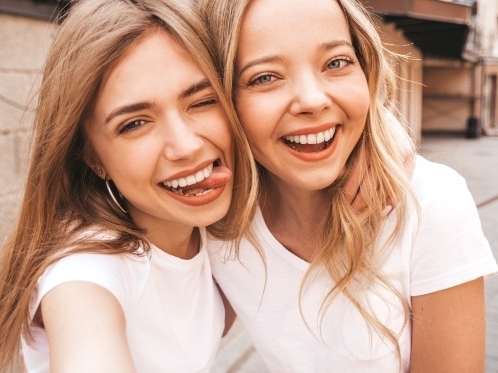 Two young smiling hipster blond women in summer white t shirt clothes. Girls taking selfie self portrait photos on smartphone.