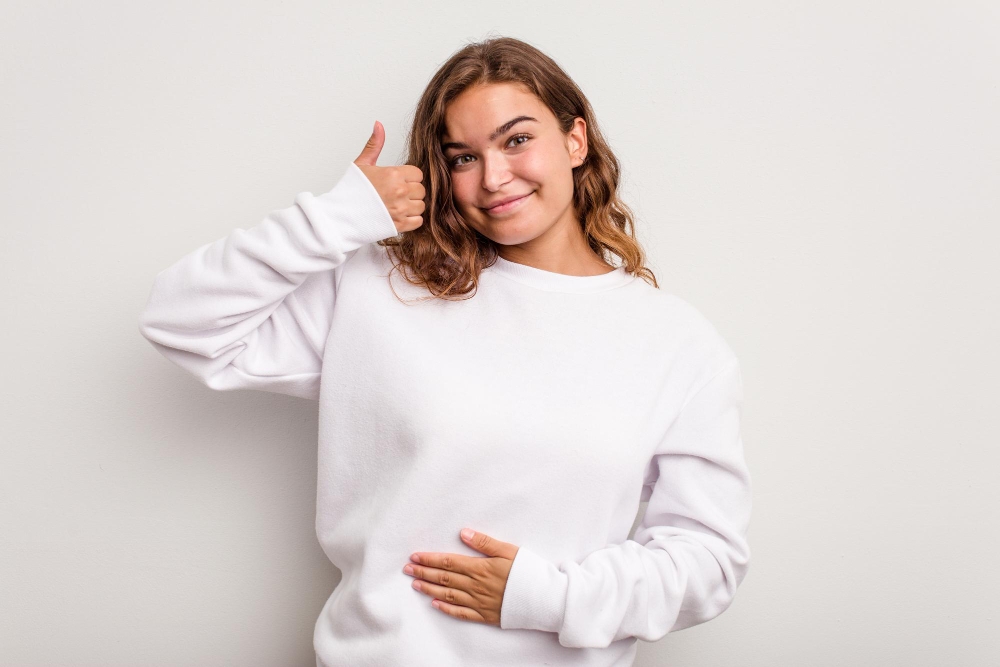 Young caucasian woman isolated on blue background touches tummy smiles gently eating
